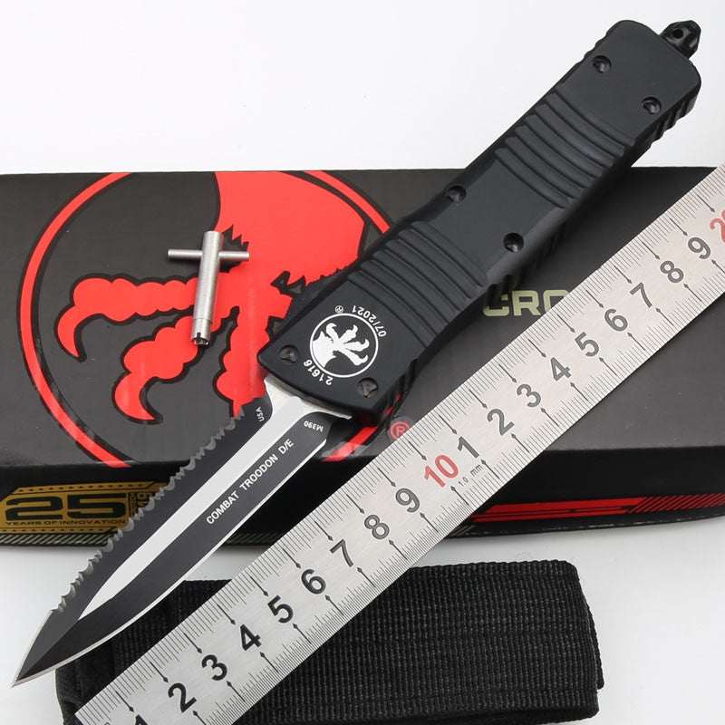 Outdoor camping tactical folding knife with black handle and serrated blade next to a ruler and nylon sheath