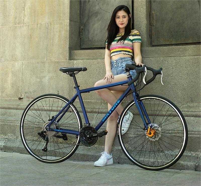 woman standing next to blue aluminum alloy road bike with flat handlebar against stone wall background