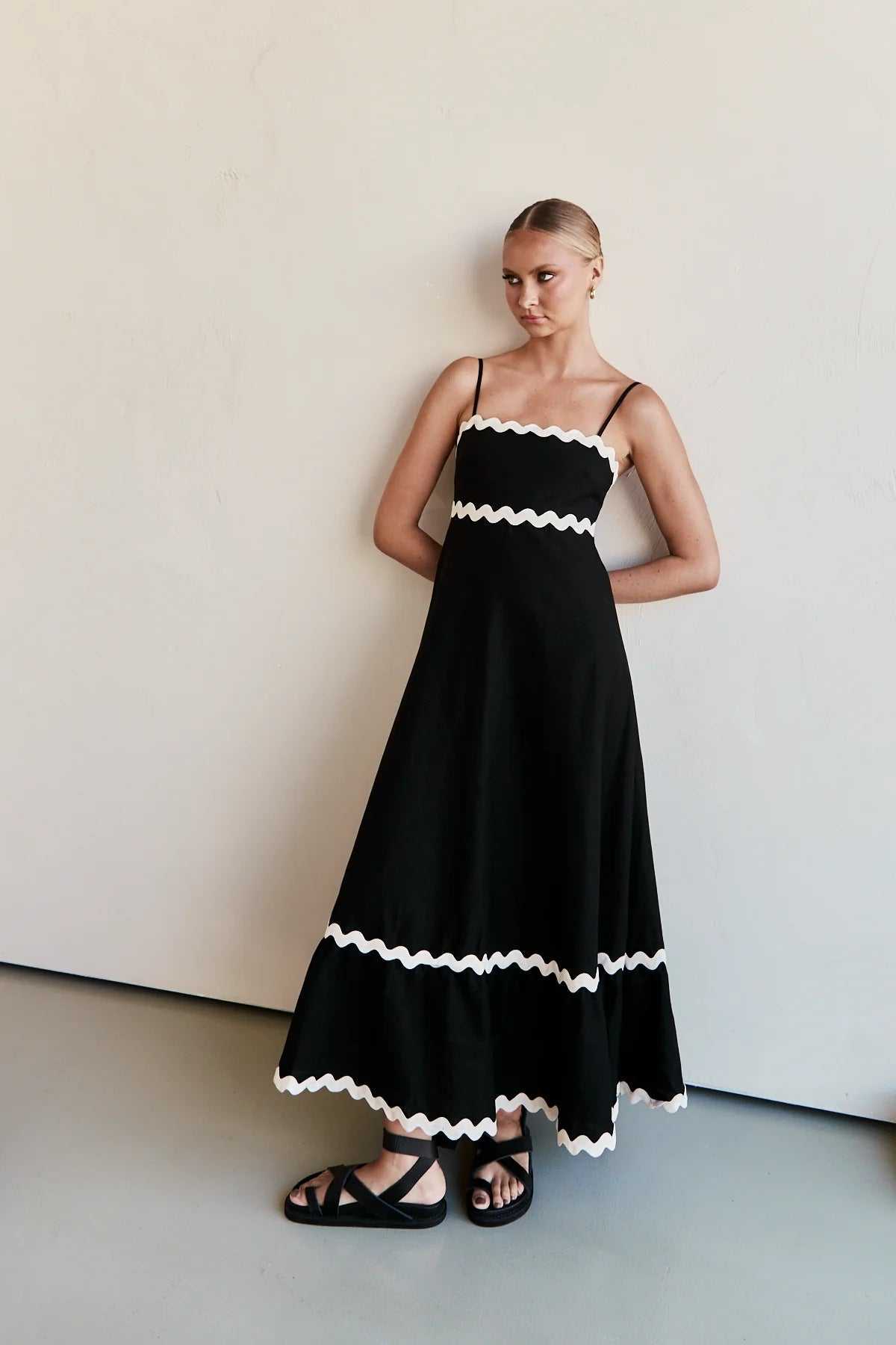 Woman wearing black suspender long dress with white wave pattern trim standing against plain wall