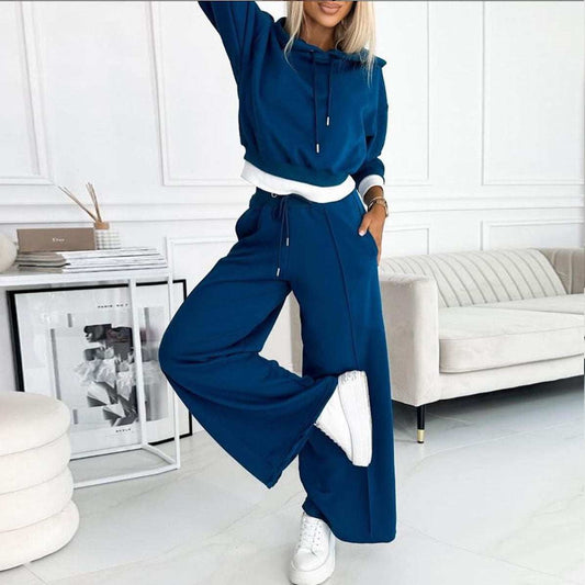 Woman wearing blue women's patchwork hooded sweater two-piece set with wide-leg trousers and white sneakers in modern living room